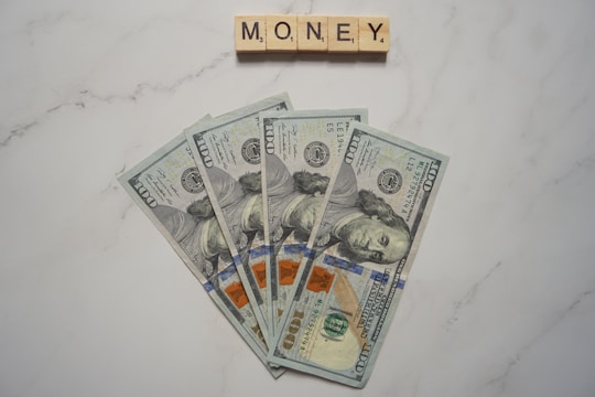 Five one-hundred dollar bills are fanned out on a marble surface. Above them, Scrabble tiles spell out the word 'MONEY'.