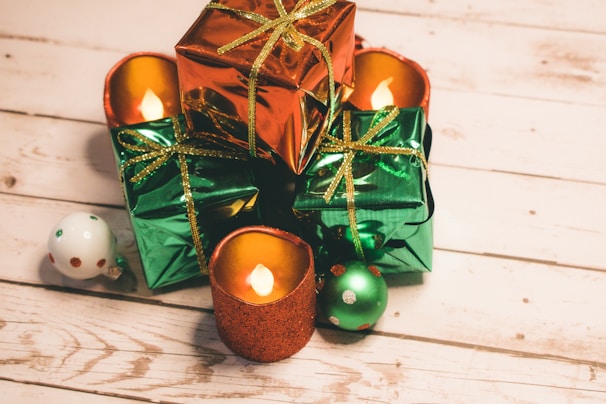 A collection of beautifully wrapped gifts in shiny red and green paper, adorned with golden ribbons, placed on a rustic wooden surface. Among the gifts are glittery candles flickering gently, creating a warm, festive atmosphere. Nearby, a couple of Christmas ornaments in coordinating colors add to the holiday decoration.