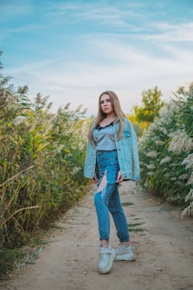 A young person stands confidently on a dirt path surrounded by tall green foliage. They wear a denim jacket over a gray top and denim jeans with a stylish rip. Their feet are adorned with white sneakers, and their long hair falls naturally. The sky is bright and clear with soft clouds visible in the background.