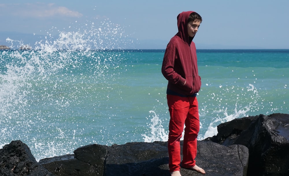 Mann in rotem Kapuzenpullover steht tagsüber auf Felsen in der Nähe des Meeres