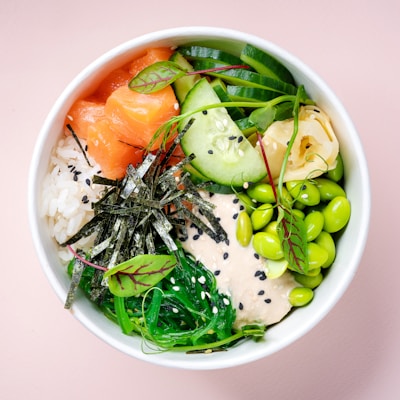 A bowl featuring a colorful assortment of fresh ingredients. There are thick slices of raw salmon, white rice topped with black sesame seeds, vibrant green edamame beans, thinly sliced cucumber, and seaweed strips. The bowl also contains creamy sauce, ginger slices, and a sprinkle of microgreens.
