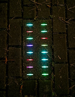 Colorful neon lights embedded in a dark pavement, arranged in neat rows. The lights glow in shades of blue, green, red, and purple, contrasting sharply with the dark background of the bricks.