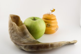 A shofar, a green apple, and a honey jar with a bee figurine on top are arranged against a plain background.