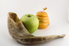 A shofar, a green apple, and a honey jar with a bee figurine on top are arranged against a plain background.