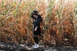A person wearing a black coat, black hat, and white boots stands in front of a field of corn plants with dry leaves. The corn stalks are tall and golden, indicating harvest season.