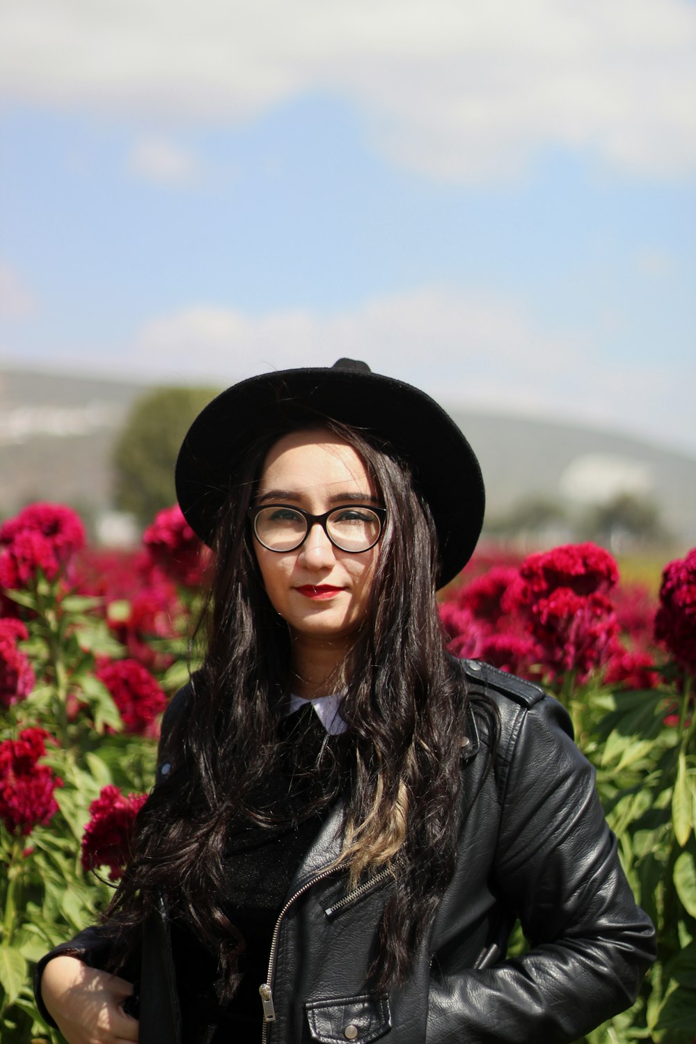 黒い帽子をかぶった黒い革ジャンを着た女性が昼間、赤い花のそばに立つ