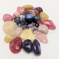 Various polished gemstones are scattered on a plain white surface, showcasing a range of colors including pink, blue, purple, green, and yellow. The stones vary in size and shape, featuring a glossy and smooth finish that reflects light.
