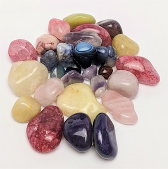 Various polished gemstones are scattered on a plain white surface, showcasing a range of colors including pink, blue, purple, green, and yellow. The stones vary in size and shape, featuring a glossy and smooth finish that reflects light.