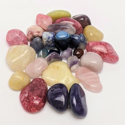 Various polished gemstones are scattered on a plain white surface, showcasing a range of colors including pink, blue, purple, green, and yellow. The stones vary in size and shape, featuring a glossy and smooth finish that reflects light.
