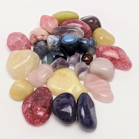 Various polished gemstones are scattered on a plain white surface, showcasing a range of colors including pink, blue, purple, green, and yellow. The stones vary in size and shape, featuring a glossy and smooth finish that reflects light.