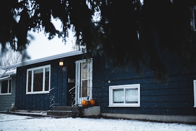 A small, cozy house is partially covered by snow, with a warm light illuminating the entrance and two pumpkins placed near the door. The house has a blue exterior and large windows, with snowflakes gently falling and visible in the ambient lighting.