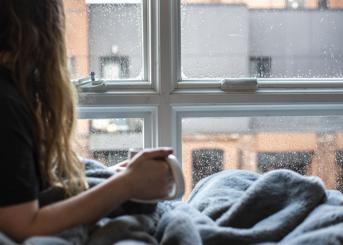 Mujer envuelta en una manta gris tomando una taza caliente junto a una ventana con gotas de condensación en un día frío de invierno