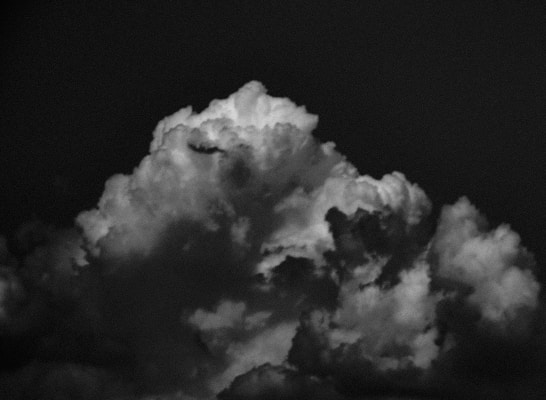 A dramatic black-and-white photograph showcasing large, billowing clouds with varying shades of gray and white, creating a striking contrast against a dark background.