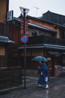 A person dressed in traditional Japanese attire walks along a street with an umbrella. The surrounding buildings feature wooden architecture typical of old Japanese houses. A street sign post and a paper lantern add to the cultural ambiance of the scene, which appears to be set on a rainy day.