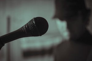 A close-up view of a microphone with a blurred person in the background, suggesting a sense of focus on the microphone itself. The setting seems to be indoors with dim lighting, creating a moody atmosphere.
