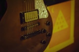 A close-up of an electric guitar focusing on the pickups, strings, and control knobs with a blurred poster in the background featuring a triangular shape and symbols.