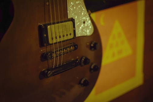 A close-up of an electric guitar focusing on the pickups, strings, and control knobs with a blurred poster in the background featuring a triangular shape and symbols.