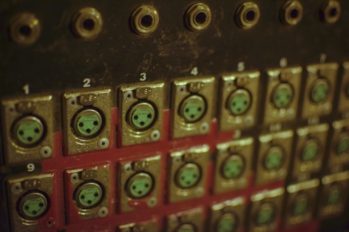 A close-up view of an old-fashioned or industrial control panel with multiple numbers, connectors, and sockets. The panel is metal, with a dark finish and some wear visible on the surface. Each connector is numbered and surrounded by a metallic frame.