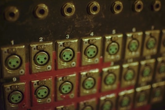 A close-up view of an old-fashioned or industrial control panel with multiple numbers, connectors, and sockets. The panel is metal, with a dark finish and some wear visible on the surface. Each connector is numbered and surrounded by a metallic frame.