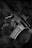 A black and white photograph depicts a close-up of a rifle resting on a tactical vest. The rifle has the phrase 'Toxic Player' written on it. The texture of the vest and metallic surface of the firearm are clearly visible.