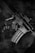 A black and white photograph depicts a close-up of a rifle resting on a tactical vest. The rifle has the phrase 'Toxic Player' written on it. The texture of the vest and metallic surface of the firearm are clearly visible.