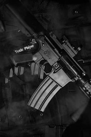 A black and white photograph depicts a close-up of a rifle resting on a tactical vest. The rifle has the phrase 'Toxic Player' written on it. The texture of the vest and metallic surface of the firearm are clearly visible.