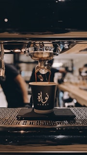 A coffee cup is positioned under an espresso machine, with coffee being dispensed into the cup. The background is a blurred image of a cafe environment, with people working and equipment visible.