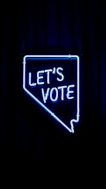 A neon sign in the shape of a state outline with the words 'LET'S VOTE' illuminated in bright blue against a dark background.