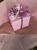 A gift box wrapped in pink paper with small black polka dots, adorned with a curly purple ribbon and a Happy Birthday tag. The box sits on top of pink tissue paper.