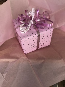 A gift box wrapped in pink paper with small black polka dots, adorned with a curly purple ribbon and a Happy Birthday tag. The box sits on top of pink tissue paper.