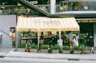 A small food stand with a yellow and white striped canopy offering Holland Fries, Beer & Snacks, and Soft Ice Cream. The stand is surrounded by various green potted plants, and several people are standing and sitting nearby. The establishment is located in front of a modern building, and a sign for 'Tiger Sugar' is visible to the right.