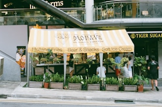 A small food stand with a yellow and white striped canopy offering Holland Fries, Beer & Snacks, and Soft Ice Cream. The stand is surrounded by various green potted plants, and several people are standing and sitting nearby. The establishment is located in front of a modern building, and a sign for 'Tiger Sugar' is visible to the right.