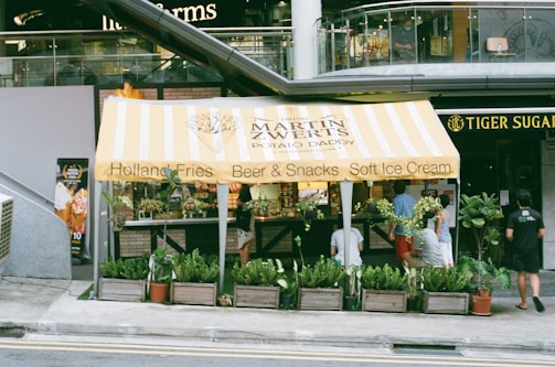 A small food stand with a yellow and white striped canopy offering Holland Fries, Beer & Snacks, and Soft Ice Cream. The stand is surrounded by various green potted plants, and several people are standing and sitting nearby. The establishment is located in front of a modern building, and a sign for 'Tiger Sugar' is visible to the right.