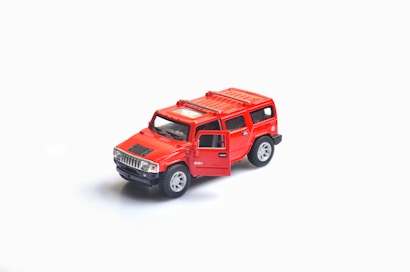 A small, detailed model of a bright red SUV with an open driver's side door, set against a plain white background. The model features silver rims and a black grille.