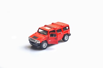 A small, detailed model of a bright red SUV with an open driver's side door, set against a plain white background. The model features silver rims and a black grille.