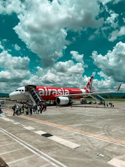 A group of people is boarding an AirAsia airplane parked on an airport tarmac under a sky filled with dramatic, fluffy clouds. The airplane is predominantly red with white lettering, and is being accessed via a boarding staircase. There are orange cones positioned around the aircraft.
