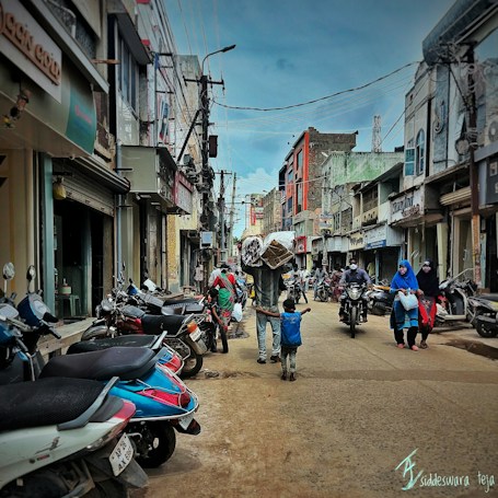 A bustling street scene with people walking and riding motorbikes. A man carrying a large bundle on his head is accompanied by a child holding his hand. Numerous parked scooters line the street, with shops on either side displaying signs in different languages.