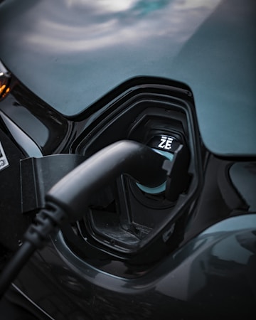 Close-up view of an electric vehicle charging port with a charging cable plugged into it. The port is labeled with the letters 'Z.E.' and is set against a smooth, dark-colored surface with a glossy finish.