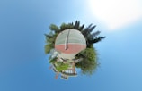 A spherical, panoramic image featuring a tennis court at its center, surrounded by trees and greenery. Buildings are visible in the lower part of the sphere, giving the impression of a miniature world with the court as its focal point. The sky forms the background, creating an expansive and open feeling.