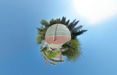 A spherical, panoramic image featuring a tennis court at its center, surrounded by trees and greenery. Buildings are visible in the lower part of the sphere, giving the impression of a miniature world with the court as its focal point. The sky forms the background, creating an expansive and open feeling.