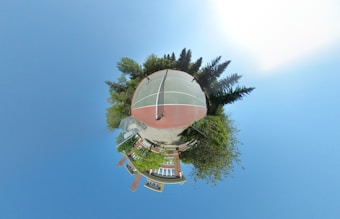 A spherical, panoramic image featuring a tennis court at its center, surrounded by trees and greenery. Buildings are visible in the lower part of the sphere, giving the impression of a miniature world with the court as its focal point. The sky forms the background, creating an expansive and open feeling.
