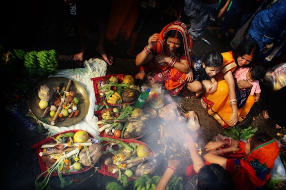 A group of women dressed in colorful traditional clothing are gathered around offerings arranged on the ground. The offerings include fruits, vegetables, and other items displayed on plates and baskets. Smoke rises in the middle, possibly from a ritual or prayer. The scene is vibrant with people actively engaged in the ritual, surrounded by bright fabrics and abundant produce.