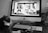 A cat is sitting in front of an Apple computer, looking at a screen displaying multiple photos of cats. The scene is in a monochrome setting, with a vase of flowers in the background and some computer peripherals on the desk.