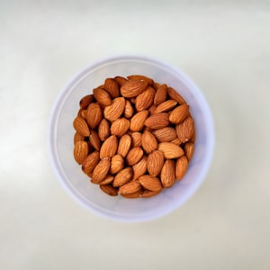 A clear plastic cup containing whole almonds is placed on a light-colored surface. The almonds are uniformly brown with a matte texture and are packed together closely within the cup.