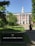A red-brick building with white columns and a central tower is surrounded by greenery and a paved path leading up to the entrance. A sign in the foreground reads 'Harvard Business School.' Several people are walking along the path towards the building.