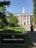 A red-brick building with white columns and a central tower is surrounded by greenery and a paved path leading up to the entrance. A sign in the foreground reads 'Harvard Business School.' Several people are walking along the path towards the building.