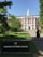 A red-brick building with white columns and a central tower is surrounded by greenery and a paved path leading up to the entrance. A sign in the foreground reads 'Harvard Business School.' Several people are walking along the path towards the building.