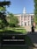 A red-brick building with white columns and a central tower is surrounded by greenery and a paved path leading up to the entrance. A sign in the foreground reads 'Harvard Business School.' Several people are walking along the path towards the building.
