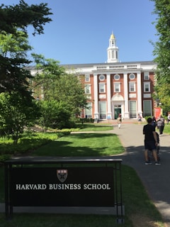 A red-brick building with white columns and a central tower is surrounded by greenery and a paved path leading up to the entrance. A sign in the foreground reads 'Harvard Business School.' Several people are walking along the path towards the building.