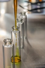 A pipette is used to fill a series of clear glass vials with a golden liquid. The vials are arranged in a line on a reflective metallic surface, likely a laboratory or industrial setting, with soft lighting creating gentle reflections on the vials.